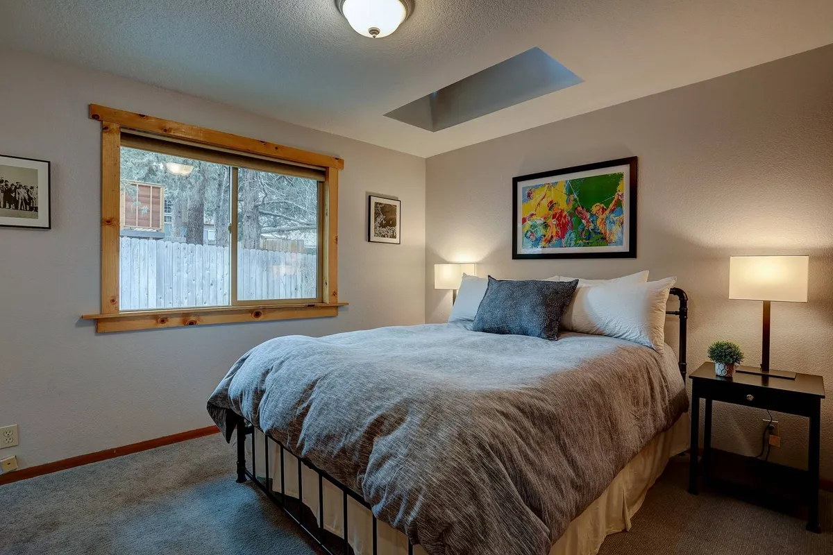 A calm, inviting bedroom featuring a cozy bed with gray bedding, a large window letting in natural light, a piece of artwork on the wall, and a nightstand with a lamp. The ambiance suggests a perfect setting for unwinding and relaxing.