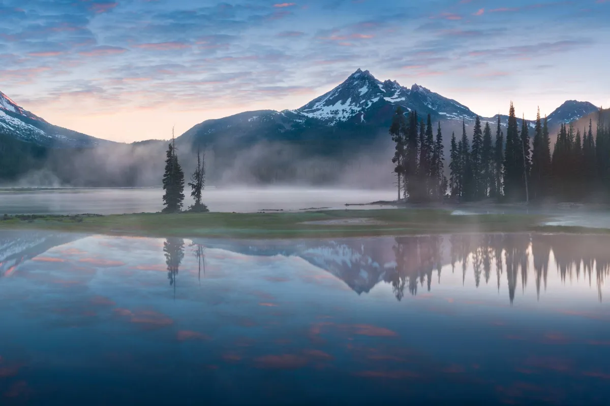 A tranquil scene of mountains reflecting on a serene lake, surrounded by lush trees and an open sky. The landscape suggests opportunities for hiking, nature photography, and peaceful reflection in all seasons.