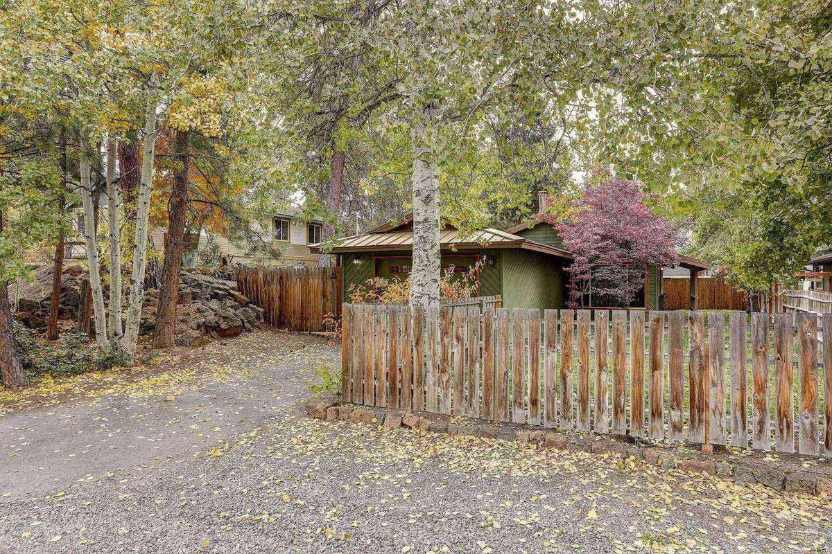 A wide view of the fenced backyard featuring a charming green house, wooden fence, and colorful trees. The scene suggests privacy and tranquility, with enough open space for outdoor games like cornhole or family gatherings. The vibrant foliage further enhances the backyard’s inviting atmosphere.