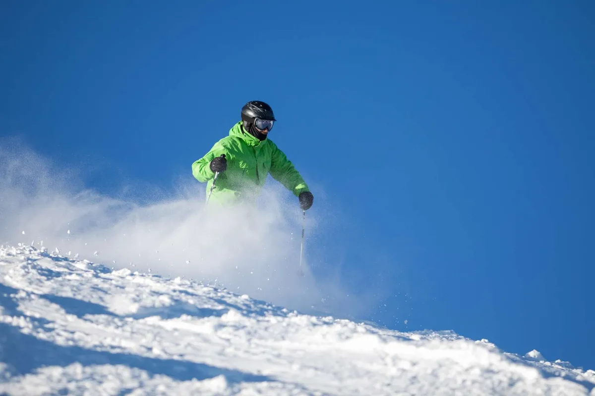 A skier in a bright green jacket carves through fresh powder on a snow-covered mountain slope beneath a clear blue sky, snow spraying in their wake. The scene is vibrant, action-packed, and filled with the thrill of winter mountain sports.
