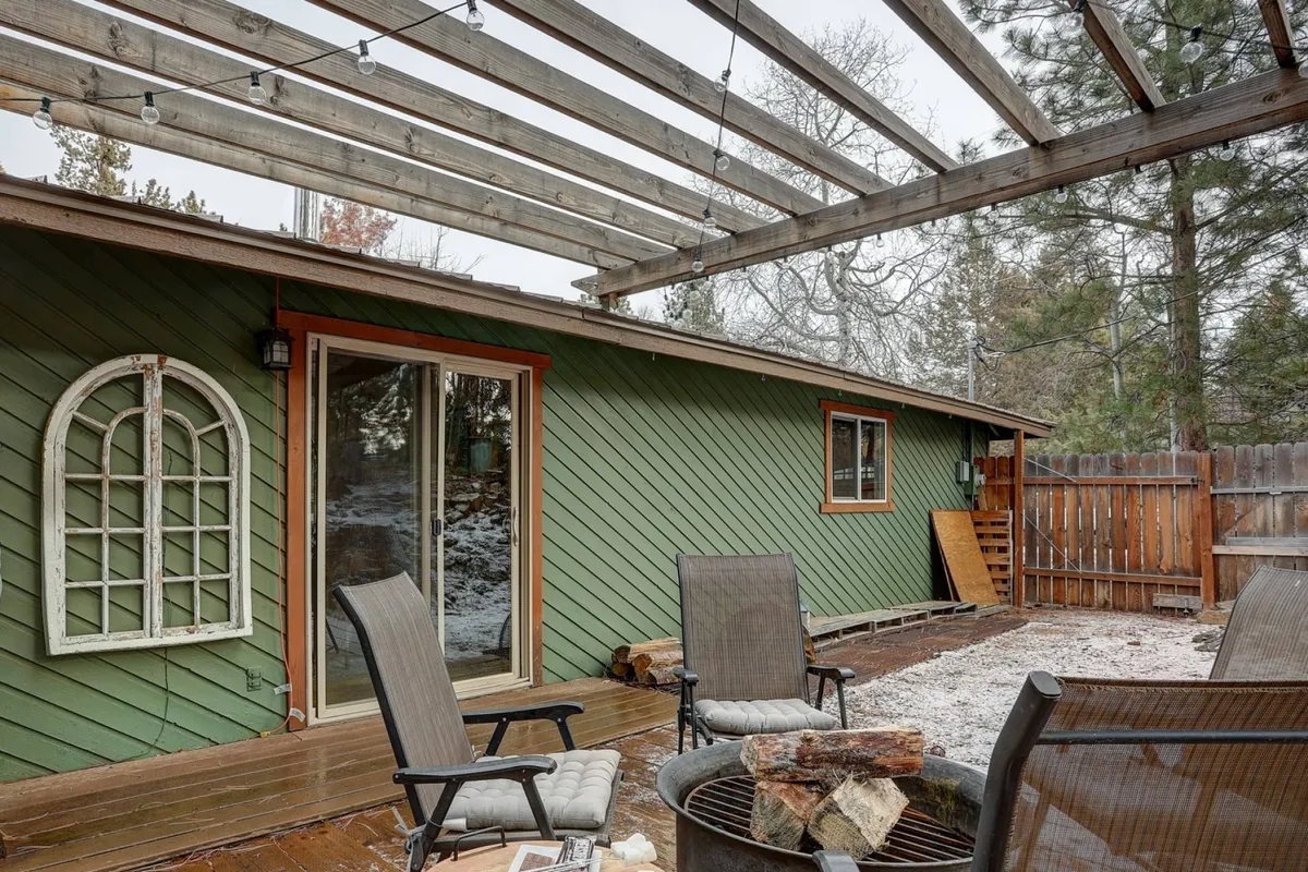 This image features a fire pit surrounded by comfortable seating, set beneath sturdy wooden beams that give the patio structure. The cozy arrangement invites groups to gather, socialize, and relax in a private backyard environment. The image is taken at an angle that showcases both the fire pit and the communal area, suggesting warmth and togetherness.
