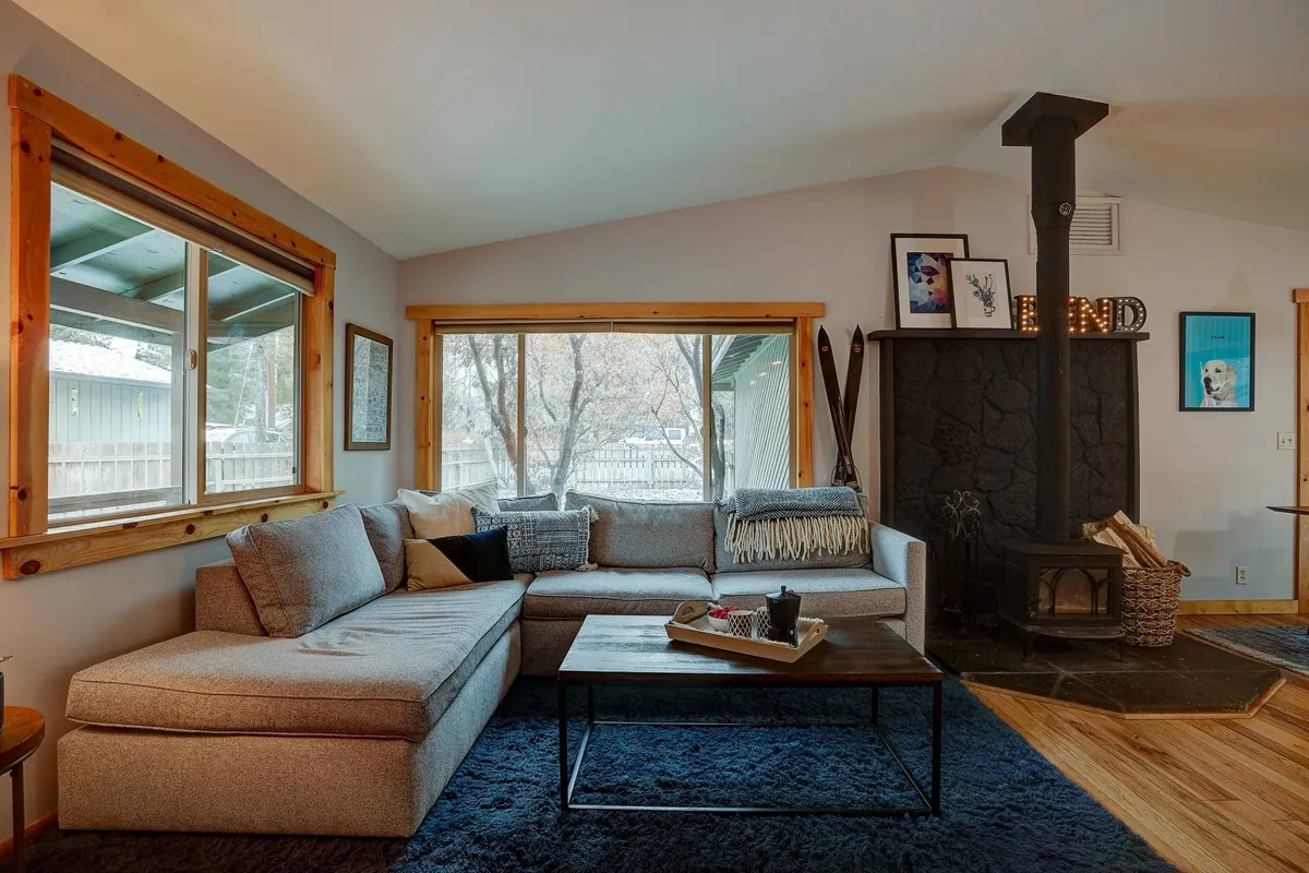 The hero image shows a spacious, sunlit living room featuring a large, comfortable sectional sofa centered around a cozy wood-burning stove. Expansive windows bring in natural light and views, while the inviting space is perfect for relaxing, socializing, or unwinding with a good book and a warm drink.