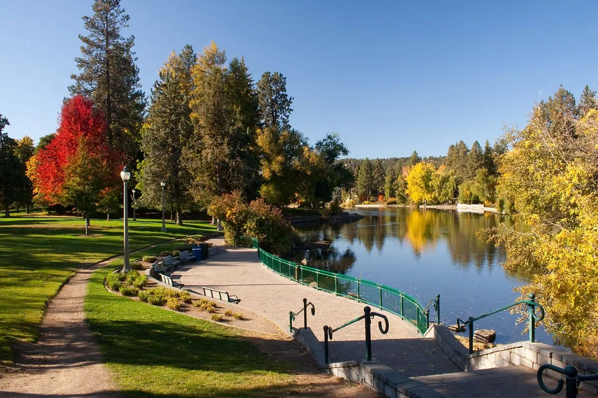 A serene lakeside setting features a scenic walking path, benches, and a charming bridge framed by trees. The water reflects the tranquil sky, inviting visitors to stroll, picnic, or pause and enjoy the outdoors.