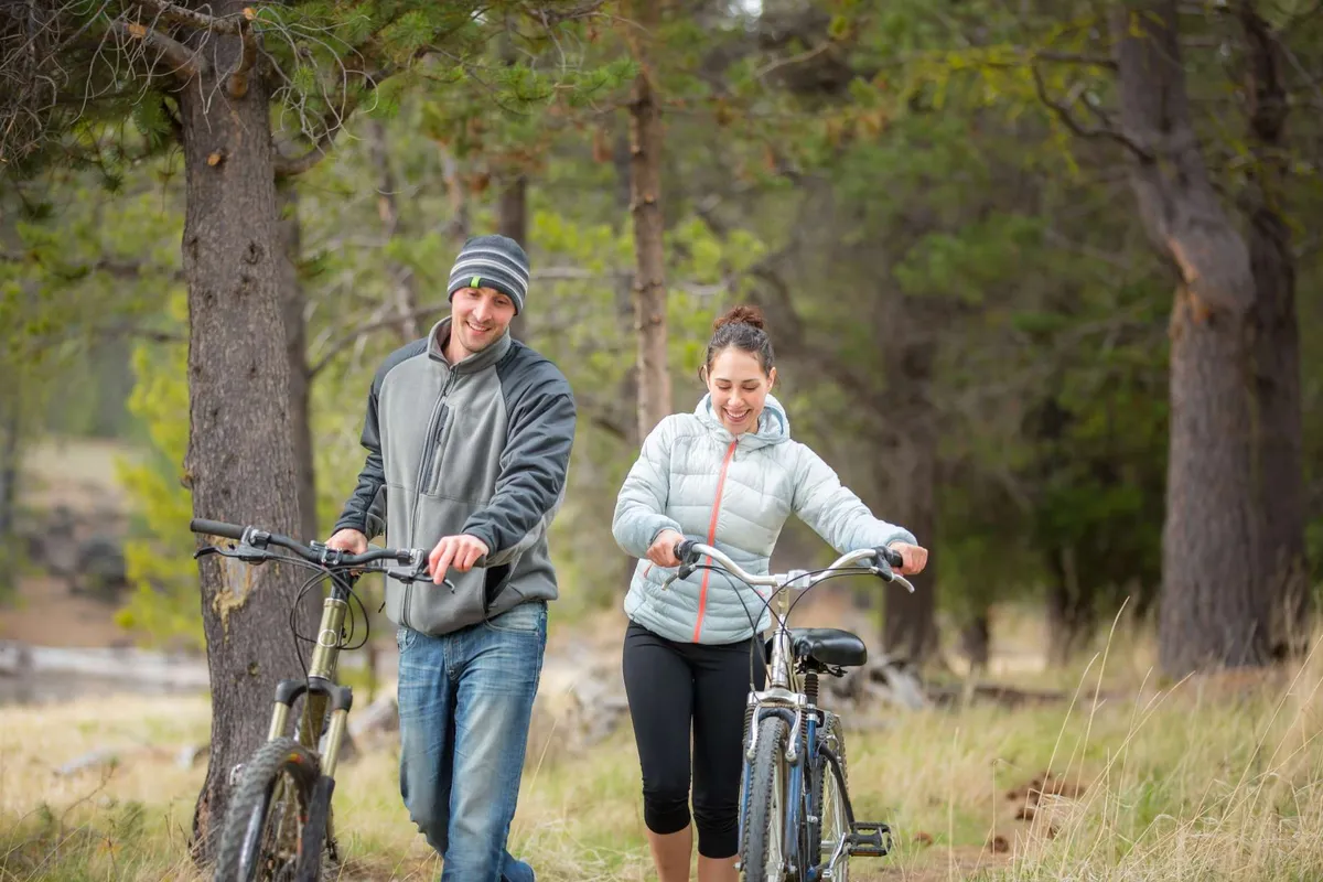 A couple walks their bicycles along a path bordered by tall trees, immersed in a natural landscape. The scene feels lively and adventurous, suggesting easy access to biking and exploring nearby trails.