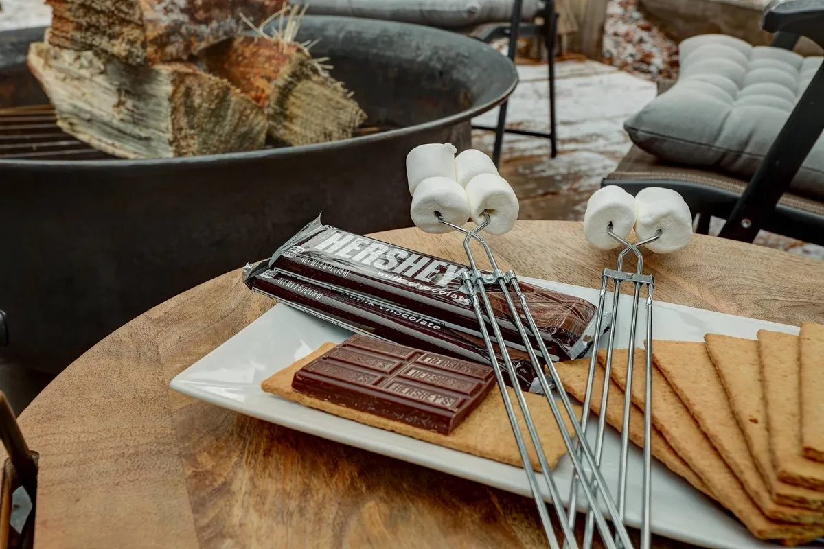 A welcoming exterior scene of a fire pit, with a wooden table spread with s'mores ingredients nearby. The setup is intimate and inviting, capturing the essence of making s'mores and enjoying treats by the fire. The mood is friendly, with the scene ready for guests to partake in classic backyard fun.