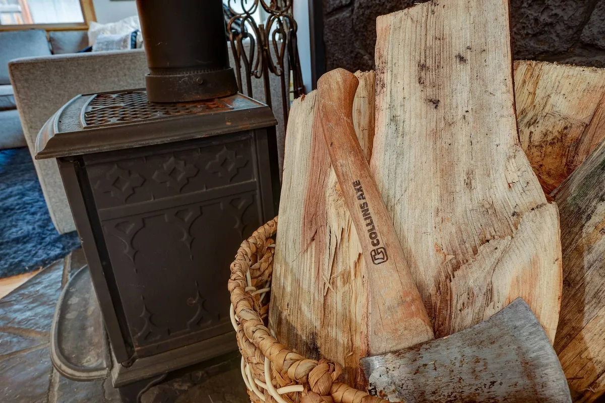 A cozy interior scene centered on a wood stove, with a tidy stack of firewood and an axe nearby. The glow of the fire creates a warm, inviting atmosphere perfect for gathering, storytelling, and unwinding after an active day.